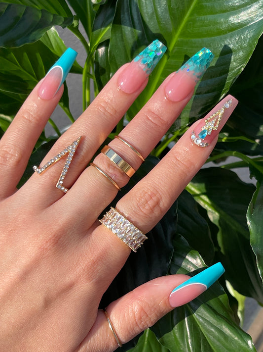 Hand with turquoise and clear nail polish wearing multiple rings against a green leafy background