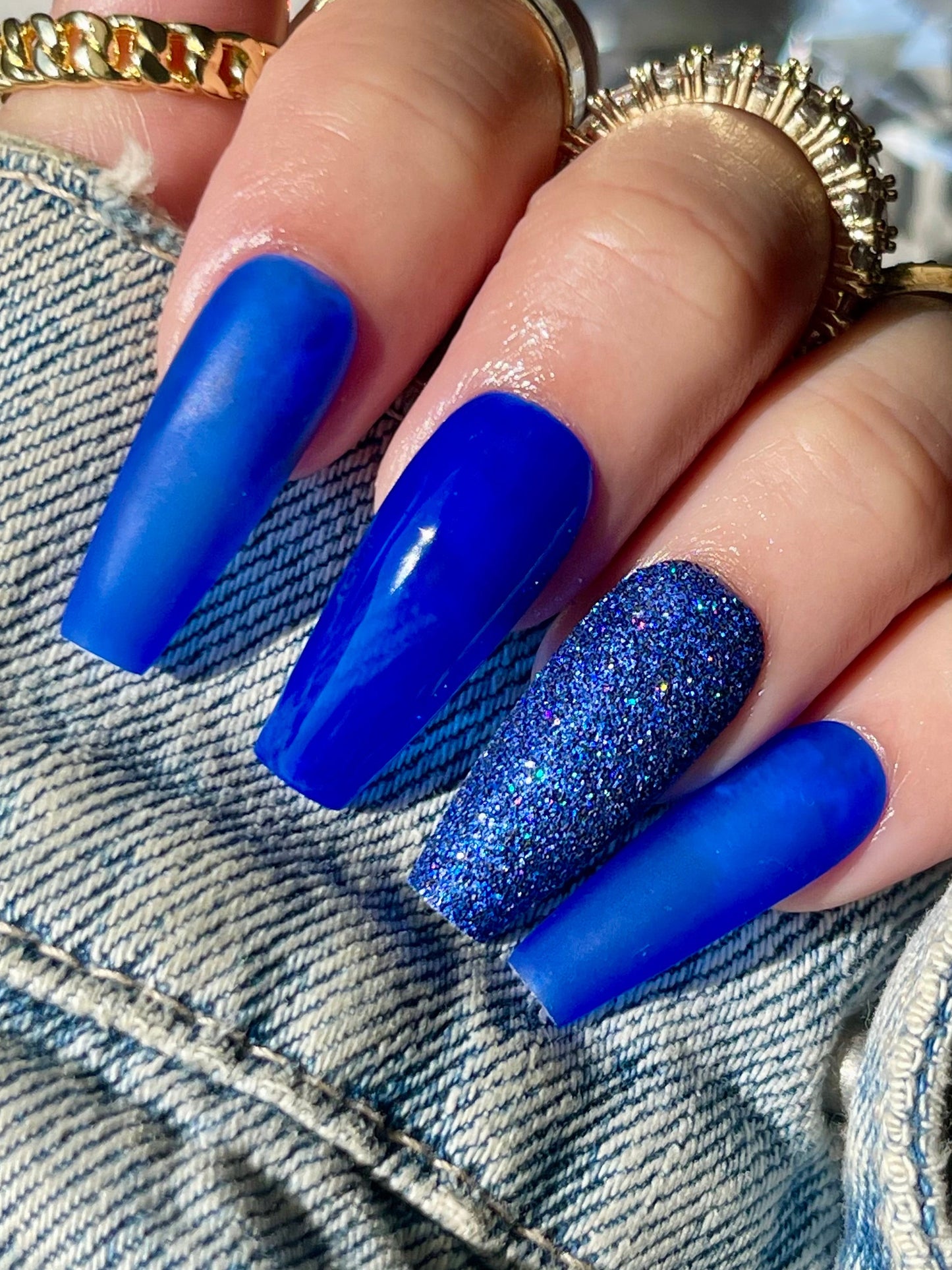 Hand with blue glittery nails wearing gold rings, denim sleeve, and crystal object in background