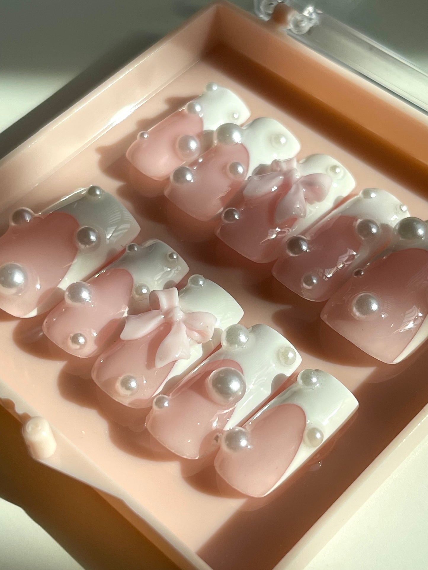 Set of pink nail art designs with pearls and bows in a display case.