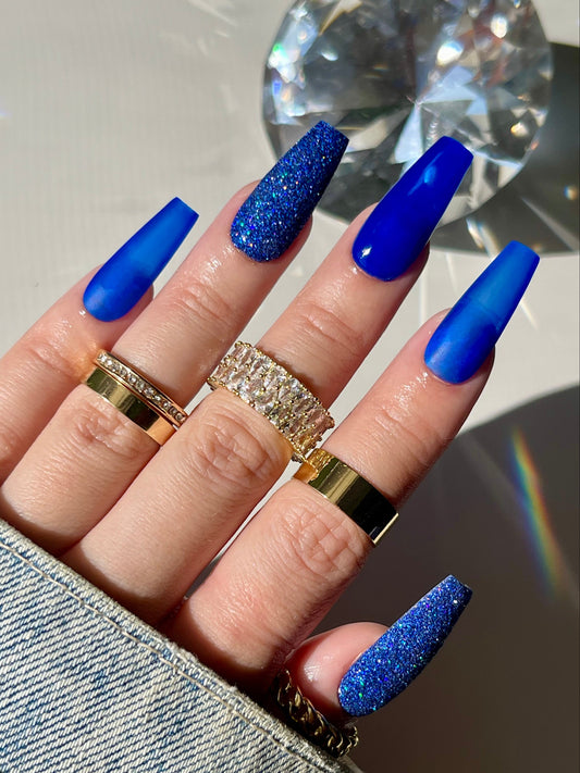 Hand with blue glittery nails and gold rings on a reflective surface
