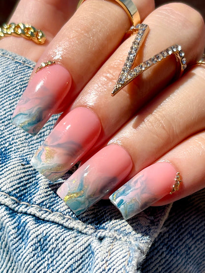 Close-up of a hand with marbled nail design wearing gold rings on denim fabric.