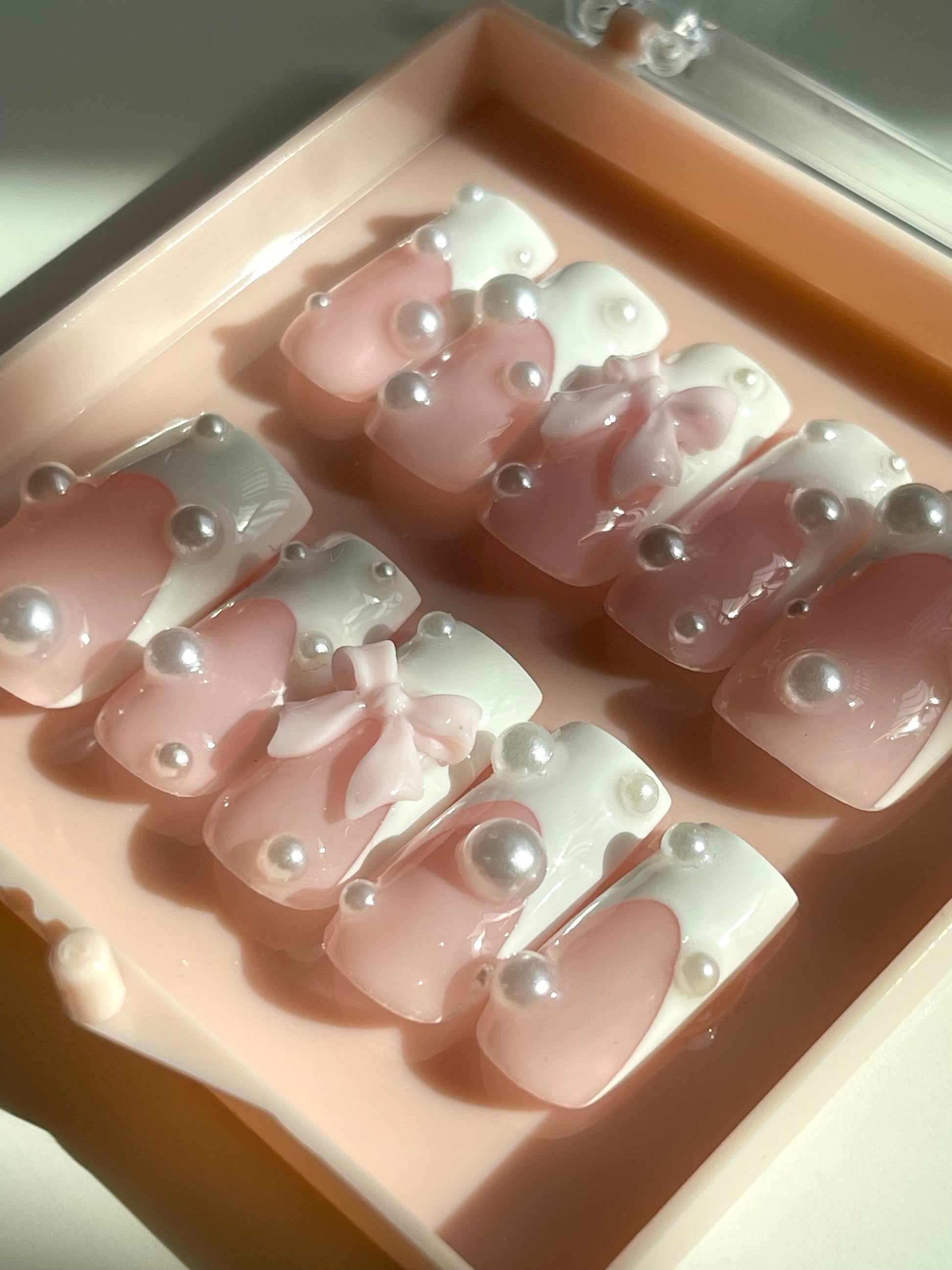 Set of pink nail art designs with pearls and bows in a display case.