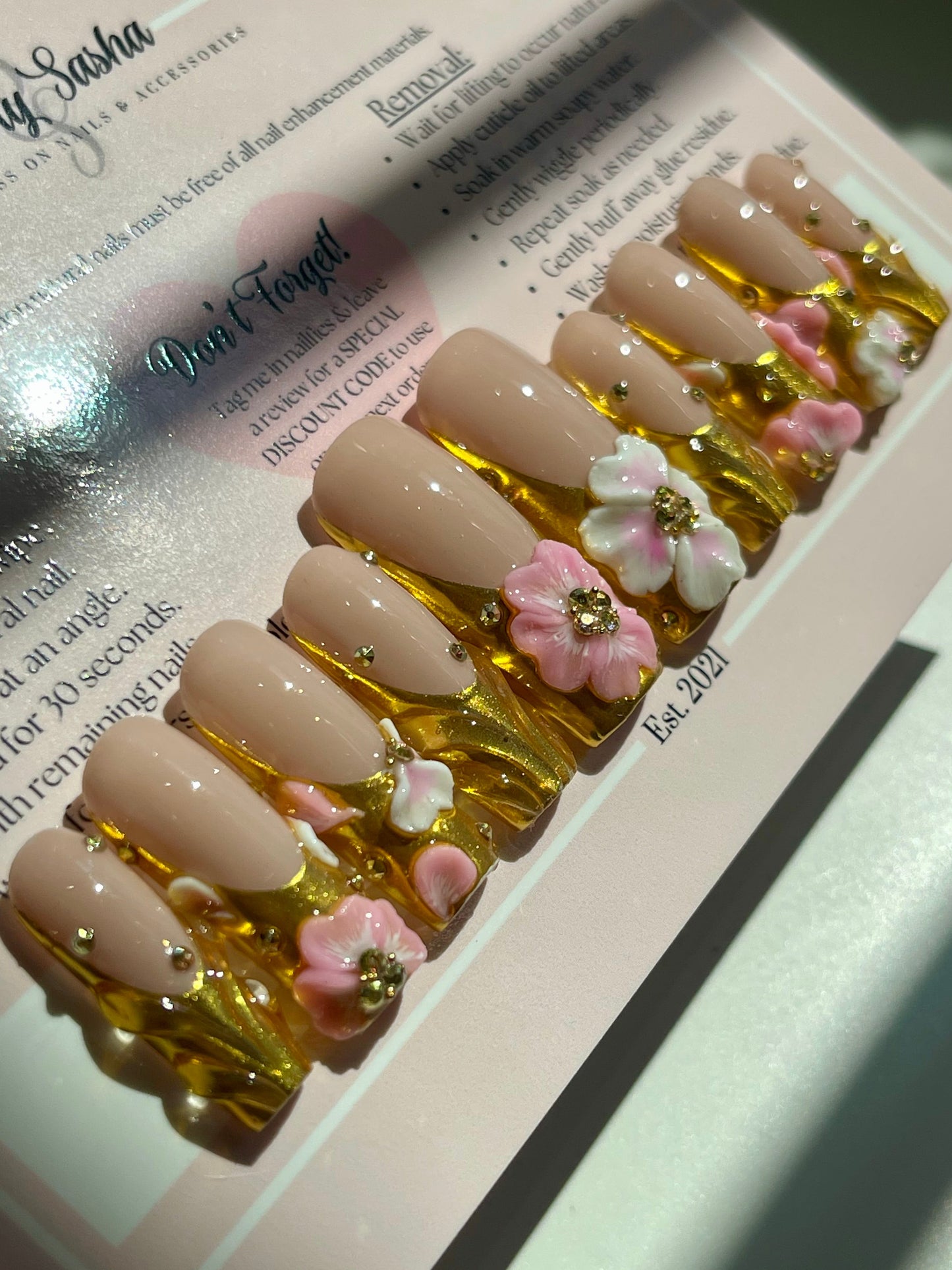 Nail art display with beige nails decorated with pink flowers and gold accents on a white surface.