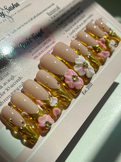 Nail art display with beige nails decorated with pink flowers and gold accents on a white surface.