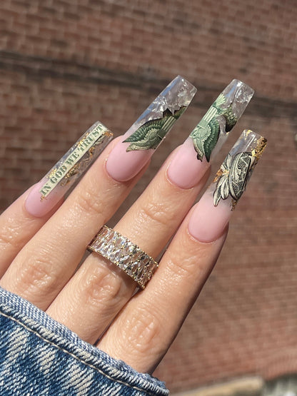 Hand wearing a diamond ring with clear nail extensions featuring dollar bill designs.