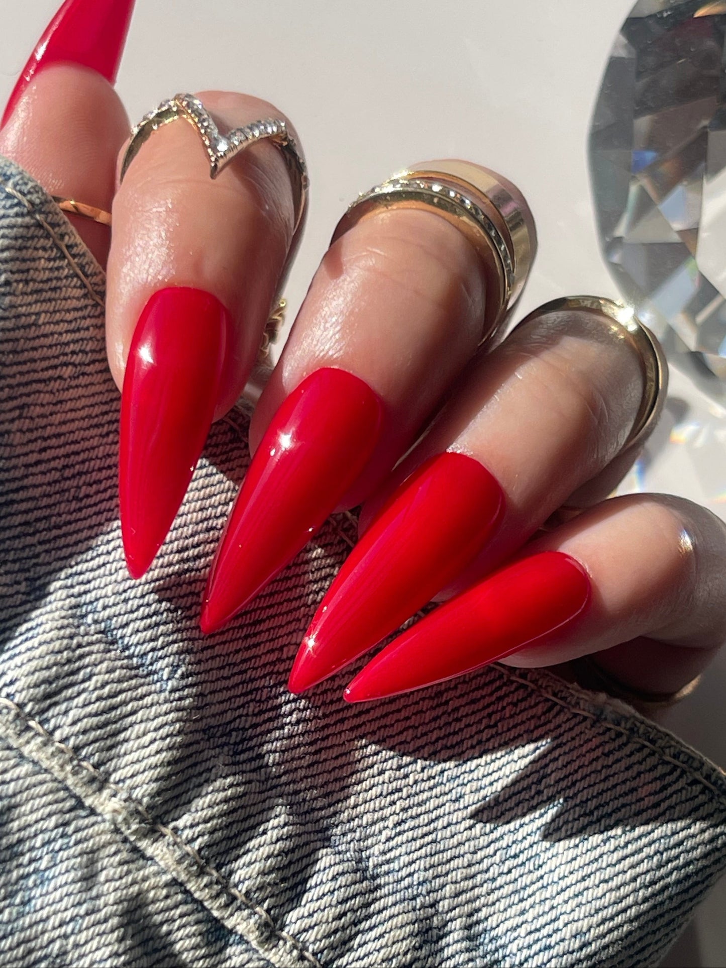 Close-up of a hand with red stiletto nails wearing gold rings on denim fabric.
