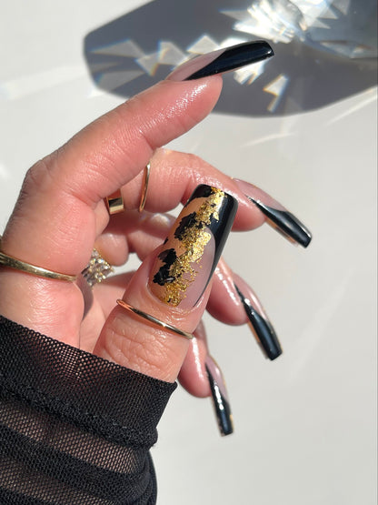 Hand with black and gold nail design wearing a gold ring on a light background